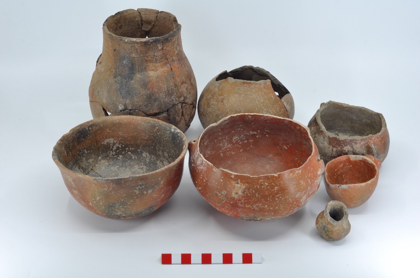 Seven early-period ceramic vessels against a grey background: jars and deep bowls, with a scale in front.